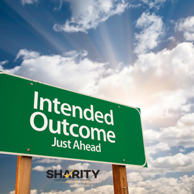 A green road sign with white text reads "Intended Outcome Just Ahead." The sign is mounted on two wooden posts and is set against a vibrant blue sky with fluffy white clouds and sun rays breaking through. In the bottom left corner, there's a logo that says "SHARITY" with the tagline "PARTNER. POWER. PROPEL." below it.